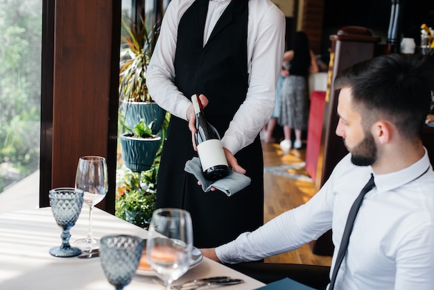 Ein junger Kellner in einer stilvollen Schürze demonstriert und bietet einem Kunden in einem Restaurant einen guten Wein an. Kundendienst.