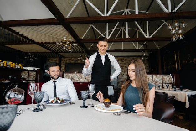 Ein junger Kellner in stilvoller Schürze serviert einen Tisch mit einem schönen Paar in einem eleganten Restaurant. Kundendienst.