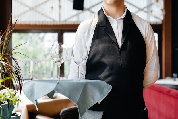 Ein junger Kellner in stilvoller Uniform steht mit Gläsern auf einem Tablett neben dem Tisch in einem schönen Gourmetrestaurant aus nächster Nähe. Restauranttätigkeit auf höchstem Niveau.