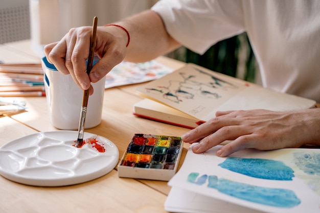 Ein junger Künstler malt mit Aquarellen in einem Notizbuch, das an einem Holztisch sitzt. Schöne männliche Hände halten eine Bürste. Nahaufnahme. Sonnenstrahlen und angenehme Atmosphäre. Bastelkonzept.Der Künstler in Aktion