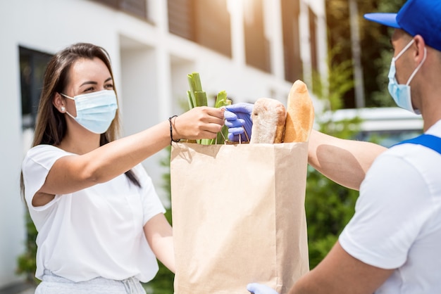 Ein junger Kurier, der eine Schutzmaske und Handschuhe trägt, liefert während der Quarantäne Waren an eine junge Frau