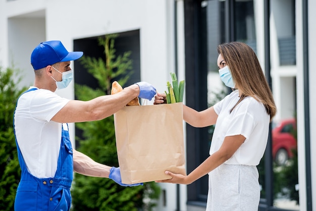 Ein junger Kurier, der eine Schutzmaske und Handschuhe trägt, liefert während der Quarantäne Waren an eine junge Frau