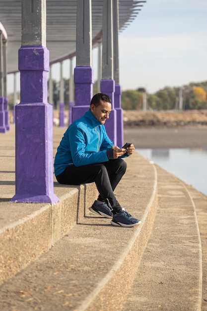 Ein junger Latino-Mann in Sportbekleidung schaut auf sein Handy, während er in einem öffentlichen Park auf einer Treppe sitzt