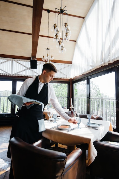 Ein junger männlicher Kellner in einer stilvollen Uniform ist damit beschäftigt, den Tisch in einem schönen Gourmetrestaurant zu bedienen. Restauranttätigkeit auf höchstem Niveau