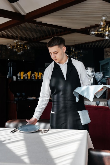 Ein junger männlicher Kellner in einer stilvollen Uniform ist damit beschäftigt, den Tisch in einem schönen Gourmetrestaurant zu servieren Ein hochrangiges Restaurant Tischservice im Restaurant