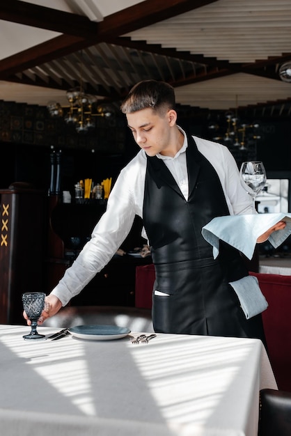 Ein junger männlicher Kellner in einer stilvollen Uniform ist damit beschäftigt, den Tisch in einem schönen Gourmetrestaurant zu servieren Ein hochrangiges Restaurant Tischservice im Restaurant