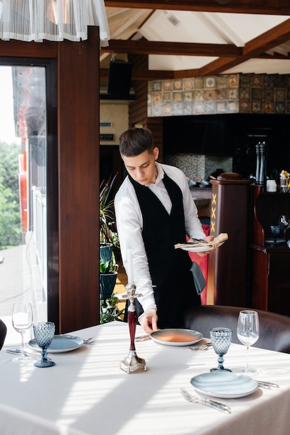Ein junger männlicher Kellner in stilvoller Uniform ist damit beschäftigt, den Tisch in einem schönen Gourmetrestaurant zu servieren. Restauranttätigkeit auf höchstem Niveau.