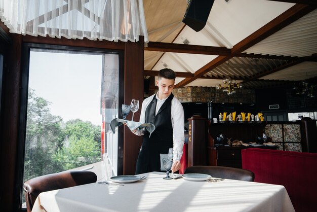 Ein junger männlicher Kellner in stilvoller Uniform ist damit beschäftigt, den Tisch in einem schönen Gourmetrestaurant zu servieren. Restauranttätigkeit auf höchstem Niveau.