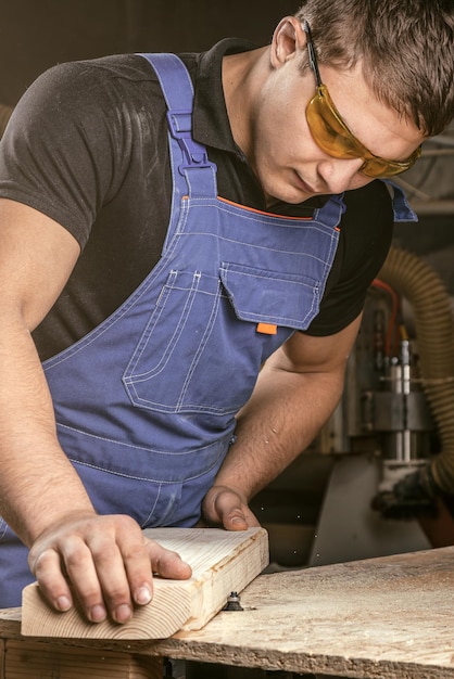 Ein junger männlicher Zimmermann in der gesamten Arbeit gleicht einer hölzernen Stange mit einer Fräsmaschine in der Werkstatt im Hintergrund