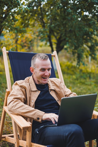 Ein junger Mann arbeitet an einem Laptop im Freien Ein junger Freiberufler ruht im Wald Fernarbeit Aktive Erholung im Sommer Tourismus Menschen Konzept Mann sitzt auf einem Stuhl im Freien