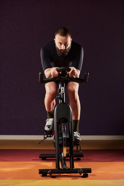 Ein junger Mann auf einem Heimtrainer mit Bart vor dunklem Hintergrund trainiert auf einem Heimtrainer im