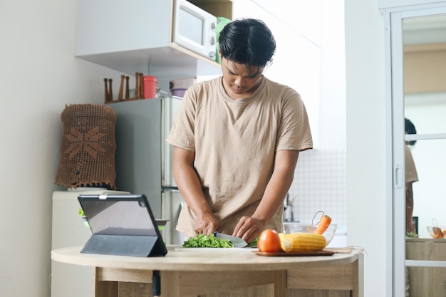 Ein junger Mann beobachtet einen Online-Kulinarkurs auf einem Tablet und macht Salat mit frischem Gemüse