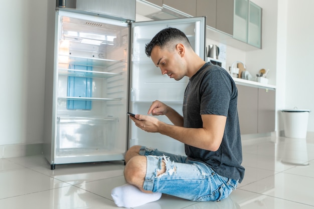 Ein junger Mann bestellt Essen mit einem Smartphone mit leerem Kühlschrank