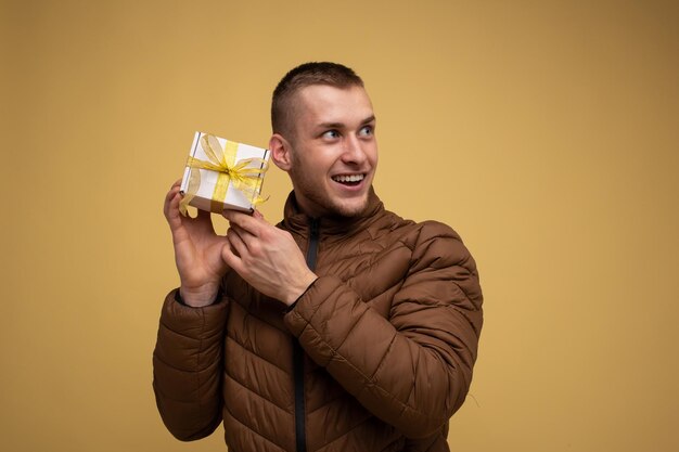 Ein junger Mann der 20er Jahre in einer braunen Jacke, auf gelbem Hintergrund, schaut zur Seite, hält sich in die Nähe des Gesichts, es gibt eine Schachtel mit einer gelben Schleife aus einem Geschenkband