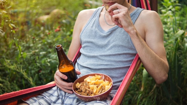 Ein junger Mann, der Bier trinkt und die Cracker stillstehen auf einer Hängematte im Sommer isst