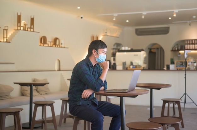 Ein junger Mann, der eine Gesichtsmaske trägt, die mit Laptop am Coffeeshop, covid-19 und Pandemiekonzept arbeitet