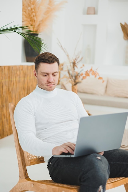 Ein junger Mann entspannt sich zu Hause, sitzt in einem bequemen Stuhl im Wohnzimmer und arbeitet an einem Laptop, während er auf seinen Laptop schaut