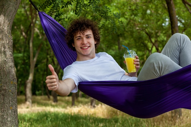 Ein junger Mann hält mit einer Hand einen Orangensaft und zeigt mit der anderen Hand Klasse, während er in einer lila Hängematte liegt und die Natur genießt