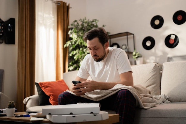 Ein junger Mann hat seine Lieblingspizza gegessen, jetzt sitzt er auf einer mit einer Decke bedeckten Couch und hält ein Telefon in der Hand