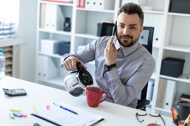 Ein junger Mann im Büro sitzt an einem Tisch, telefoniert und gießt Kaffee in eine Tasse.