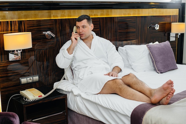 Ein junger Mann im weißen Laborkittel sitzt auf dem Bett und telefoniert in seinem Hotelzimmer.