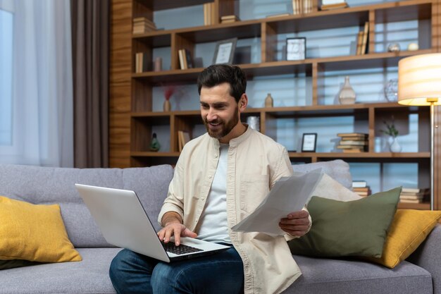 Ein junger Mann in einem beigen Hemd sitzt mit einem Laptop zu Hause auf dem Sofa und arbeitet mit Dokumentenrechnungen