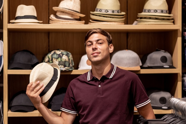 Ein junger Mann in einem burgunderfarbenen T-Shirt und einer beigen Hose mit einem Strohhut in der Hand posiert zwischen Holzregalen mit verschiedenen Dingen und Taschen in verschiedenen Farben in den Händen. Porträt. Rabatte. Verkauf.