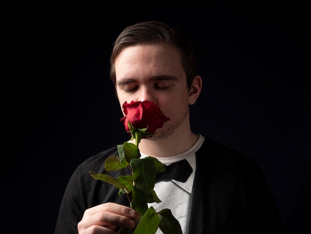Ein junger Mann in einem schwarzen T-Shirt-Anzug hält eine rote Rose in den Händen und schnüffelt mit geschlossenen Augen an Schwarz