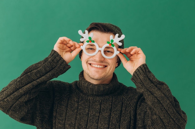 Ein junger Mann in einem warmen Pullover, einer Mütze und einer Karnevalsbrille isoliert auf grünem Hintergrund