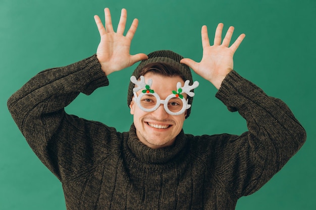 Ein junger Mann in einem warmen Pullover, einer Mütze und einer Karnevalsbrille isoliert auf grünem Hintergrund