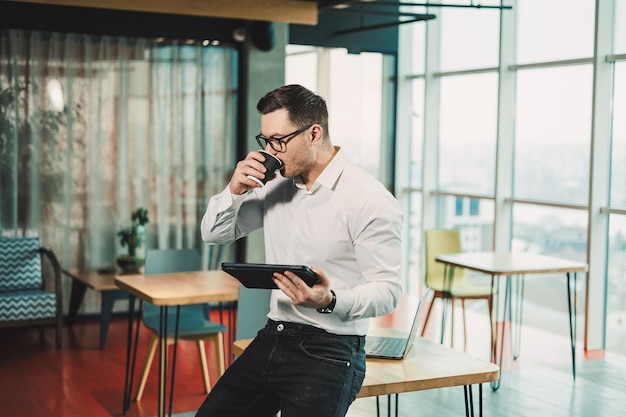 Ein junger Mann in einem weißen Hemd steht in einem geräumigen modernen Büro mit Kaffee und arbeitet an einem Tablet Moderner Büroangestellter Geschäftsmann in Brillen arbeitet online und trinkt entspannt heißen Kaffee