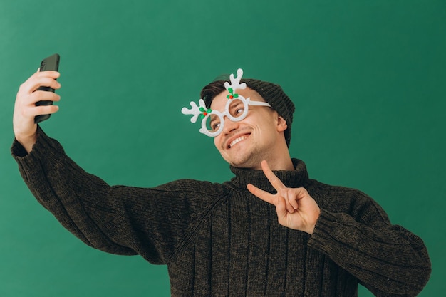 Ein junger Mann in einer warmen Pullover-Karnevalsbrille und einem Telefon in seinen Händen isoliert auf grünem Hintergrund