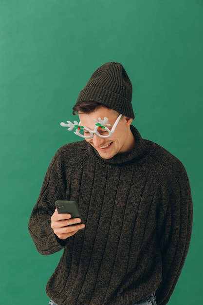 Ein junger Mann in einer warmen Pullover-Karnevalsbrille und einem Telefon in seinen Händen isoliert auf grünem Hintergrund
