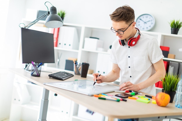 Ein junger Mann in Gläsern steht in der Nähe eines Computertisches. Ein junger Mann zeichnet einen Marker auf eine Magnettafel. Am Hals hängen die Kopfhörer des Typen.