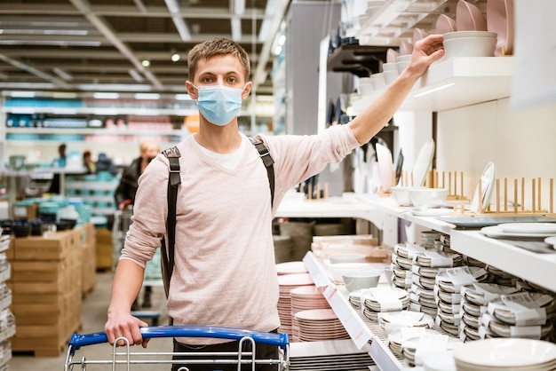 ein junger Mann, in Schutzmaske und in Freizeitkleidung, wählt das Geschirr im Laden in der Abteilung Alles für die Küche aus