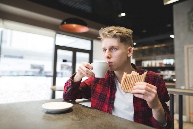 Ein junger Mann isst ein Sandwich und trinkt Kaffee in einem gemütlichen Café. Der Student spült in einem Café und schaut nachdenklich in das Fenster