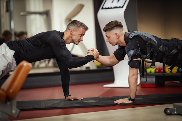Ein junger Mann macht Plank-Übungen mit Personal Trainer im Fitnessstudio, gekleidet in einen schwarzen Anzug mit einem elektronischen EMS-Simulator, um seine Muskeln zu stimulieren.