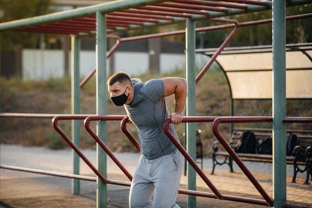 Ein junger Mann macht Push-UPS, Pull-UPS auf einem Sportplatz in einer Maske während einer Pandemie bei Sonnenuntergang. Sport, gesunder Lebensstil.