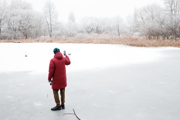 Ein junger Mann mit Bart geht im Winter auf einem zugefrorenen See spazieren und fotografiert die Natur auf seinem Handy