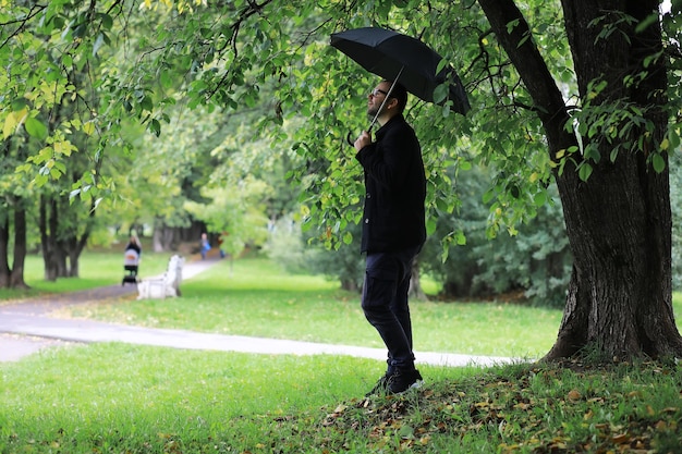 Ein junger Mann mit Brille geht während des Regens mit einem Regenschirm durch den Park.