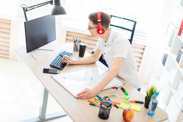 Ein junger Mann mit Brille und Kopfhörer steht neben einem Computertisch, hält einen Stift in der Hand und druckt auf der Tastatur. Vor ihm liegen eine Magnettafel und Marker.