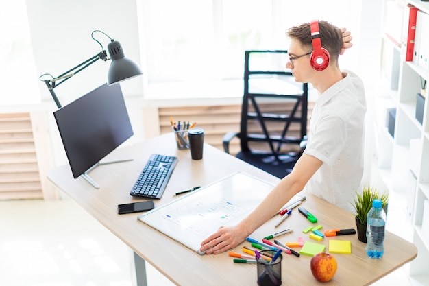 Foto ein junger mann mit brille und kopfhörer steht neben einem computertisch und kratzt sich am kopf. vor ihm liegen eine magnettafel und marker.