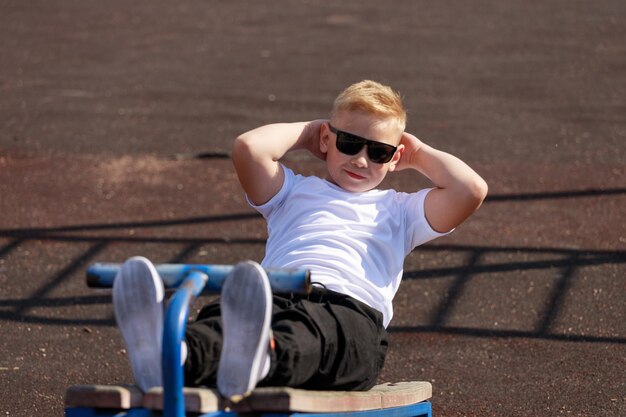 Ein junger Mann mit dunkler Brille trainiert im Sommer auf dem Sportplatz die Muskeln der Presse. Foto in hoher Qualität