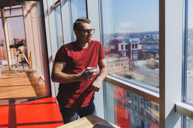 Ein junger Mann mit einer roten T-Shirt-Brille zur Sehkorrektur im Gesicht Er steht mit einer Tasse Kaffee am Fenster und blickt auf die Stadt