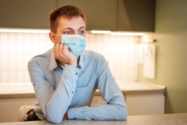 Ein junger Mann mit kaukasischem Aussehen sitzt in einem hellblauen Hemd und einer medizinischen Maske in der Küche.