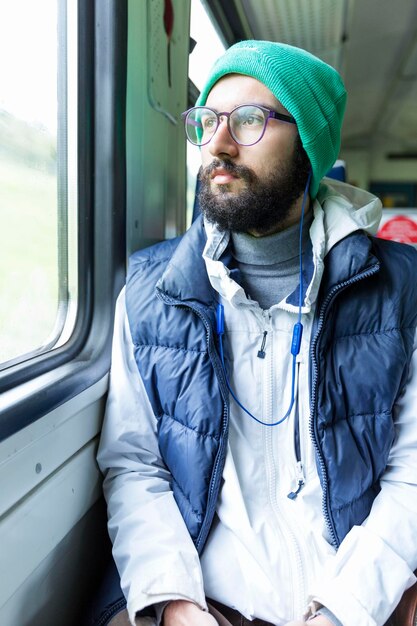 Ein junger Mann mit Kopfhörern fährt mit dem Zug und schaut aus dem Fenster Schöner ernster Mann mit Bart Vertikal