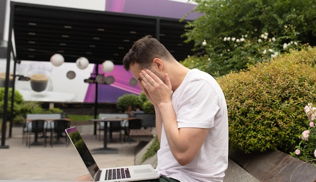 Ein junger Mann mit Kopfschmerzen sitzt im Freien und benutzt einen Laptop