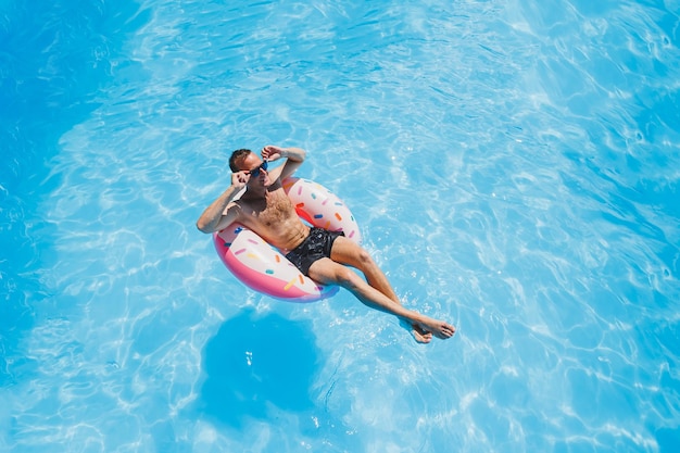 Ein junger Mann mit Sonnenbrille und Shorts entspannt sich auf einem aufblasbaren Donut im Pool Sommerurlaub