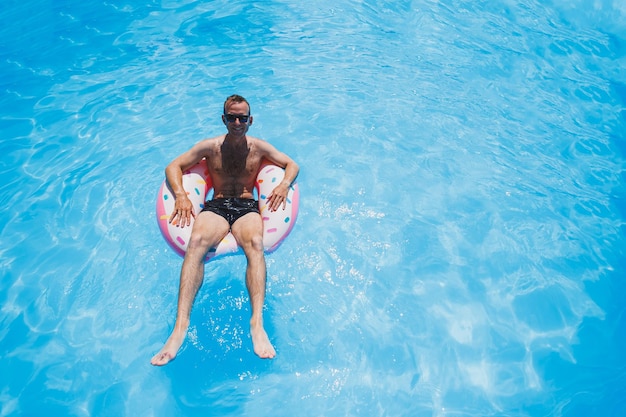 Ein junger Mann mit Sonnenbrille und Shorts entspannt sich auf einem aufblasbaren Donut im Pool Sommerurlaub