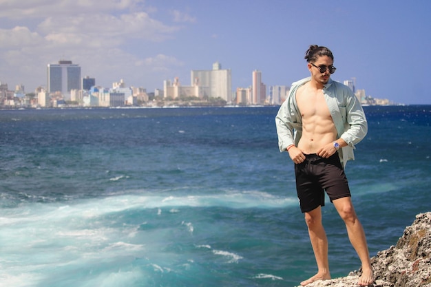 Ein junger Mann ohne Hemd steht hinter dem Meer und trägt eine weiße Hose. Er ist ein junger Latino mit Lachen